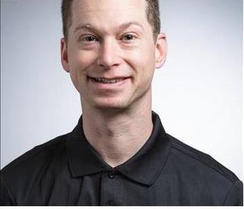 Male controller wearing black collared shirt smiling in front of grey background